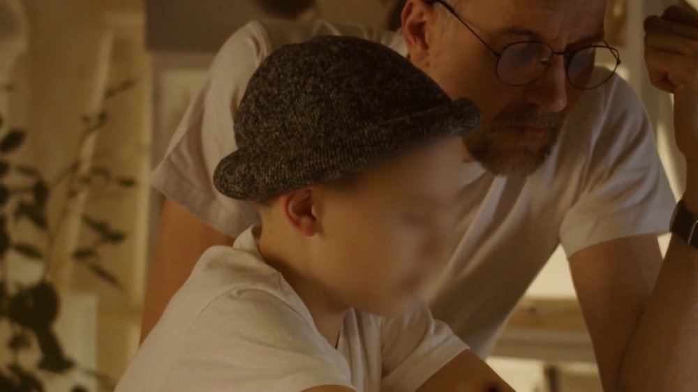 En vuxen person med glasögon lutar sig fram bredvid ett barn i grå hatt och vit skjorta. Båda verkar vara fokuserade på något framför dem. Scenen är varmt upplyst inomhus.